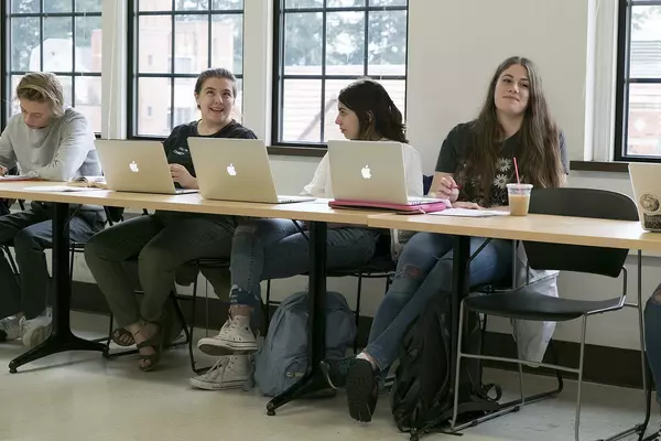 Four students sit at a long table with Apple Macbooks in front of them.