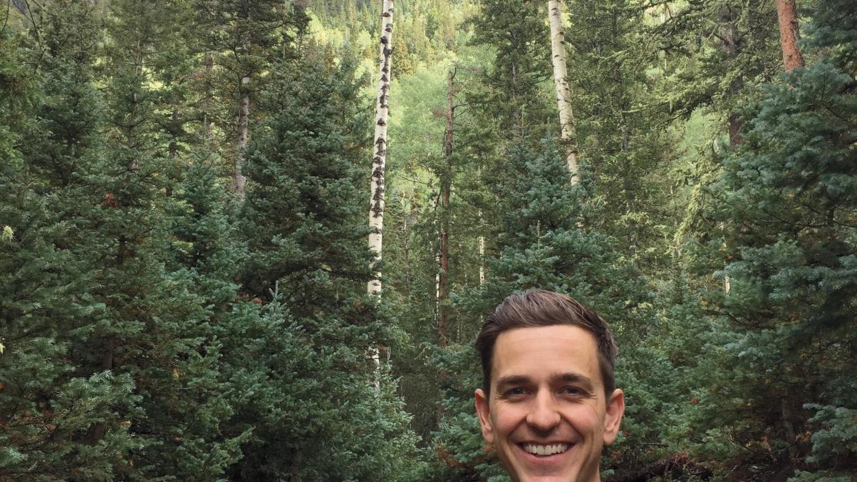 Man in a black shirt smiling with a forest in the background