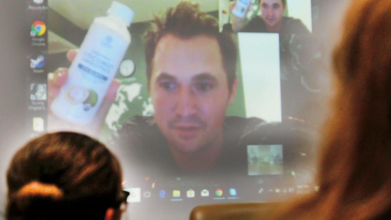 A person holding a bottle of coconut water, presenting to people from a computer screen