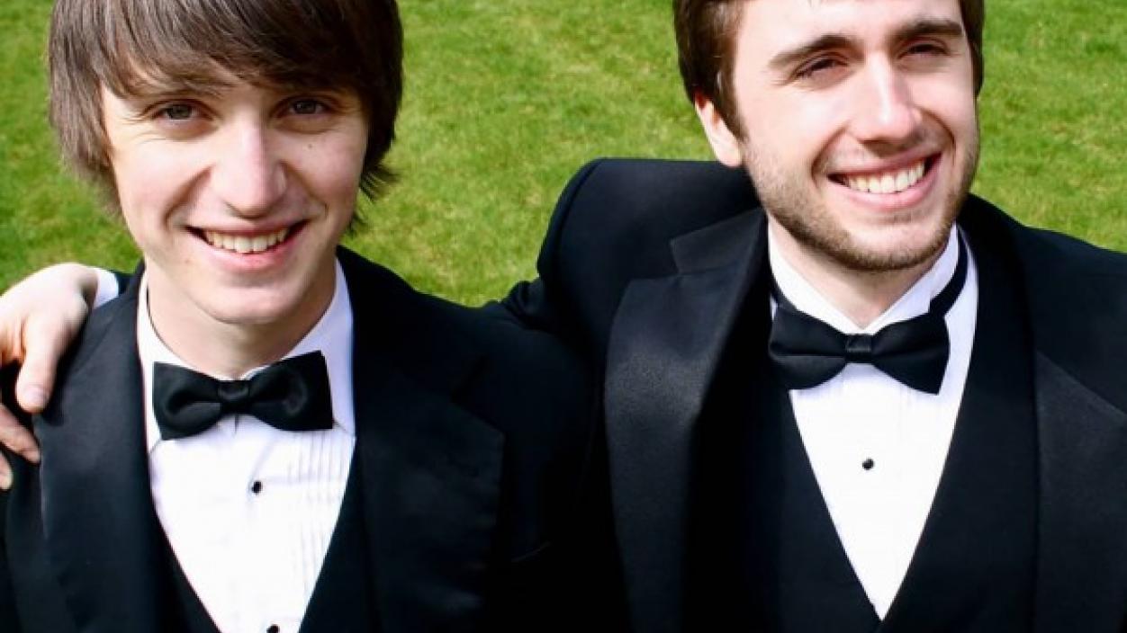 Two people in tuxedos smiling and standing outside