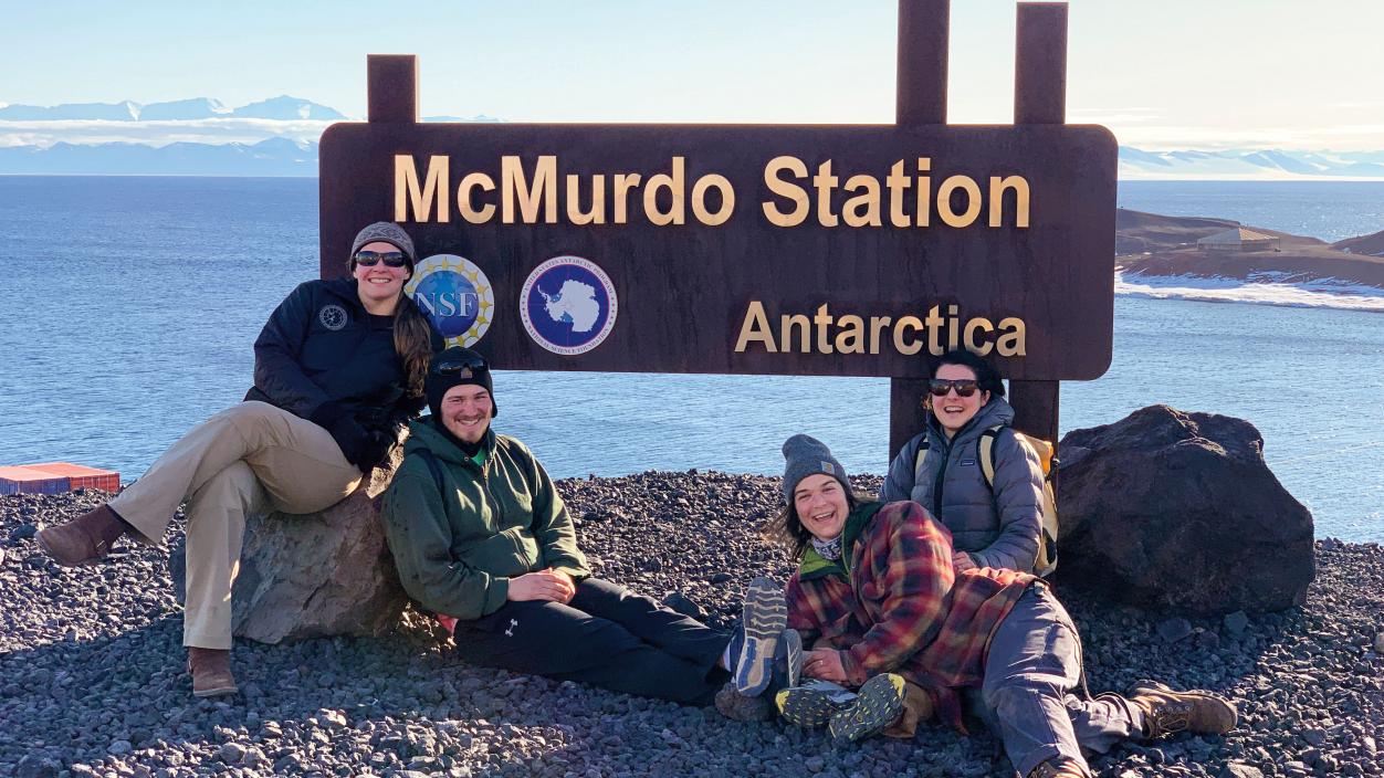 Four people posing with a McMurdo Station signpost in Antarctica