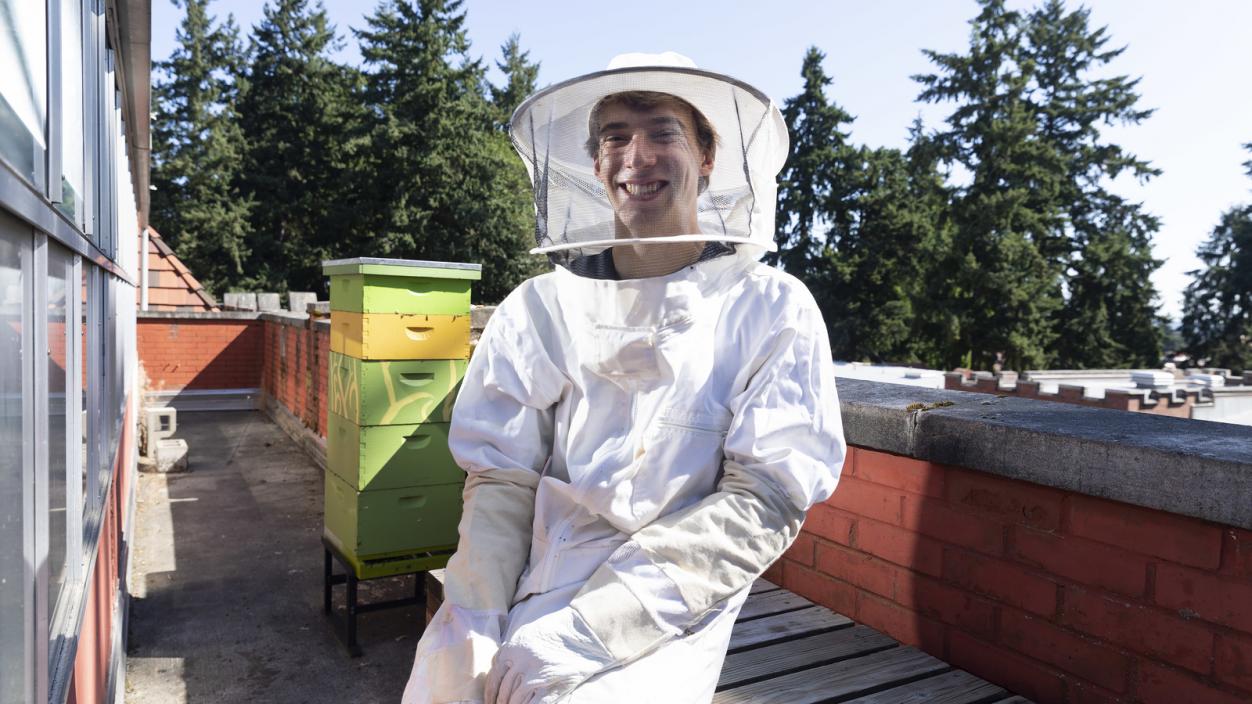 Adam Schmidt ’23, dressed in a protective suit, stands near Puget Sound's bee hives on the roof of Thompson Hall
