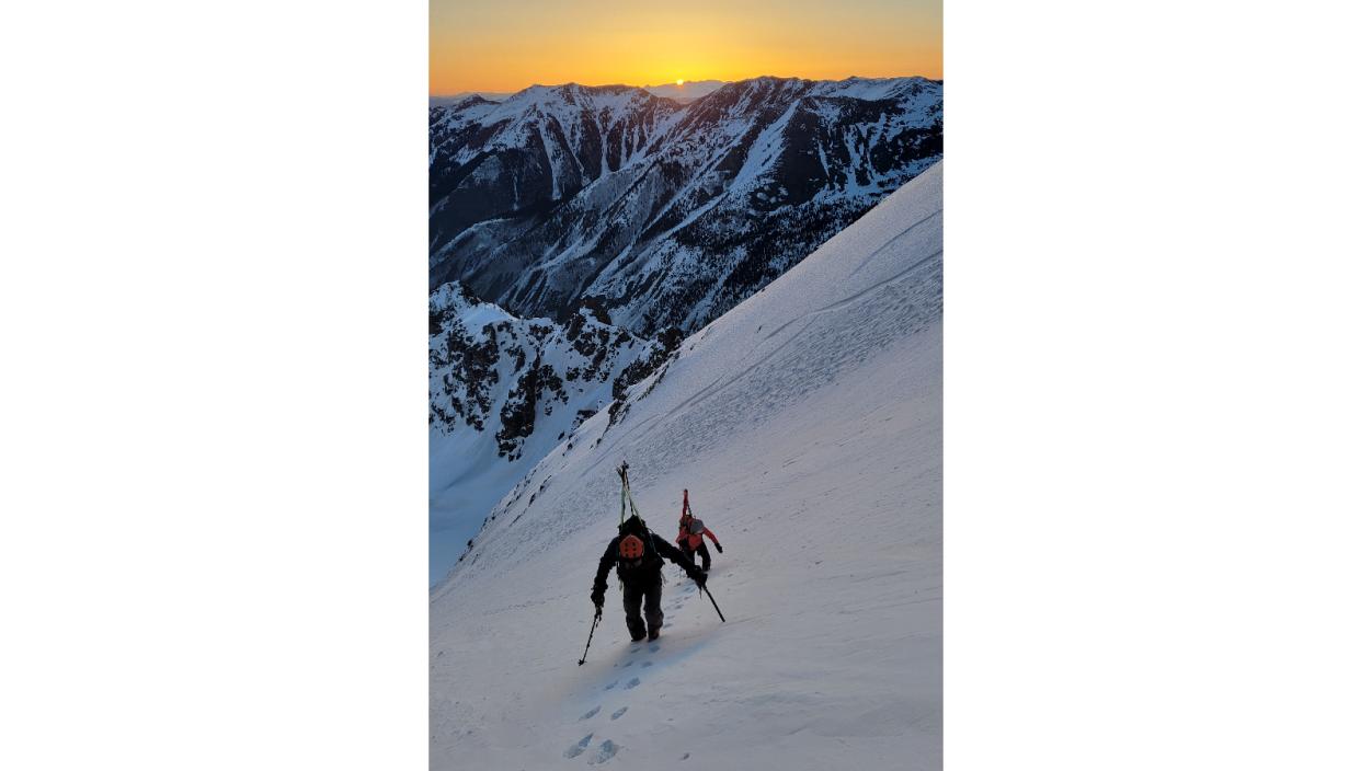 Two climbers ascend a snowy embankment with the rising sun behind them.