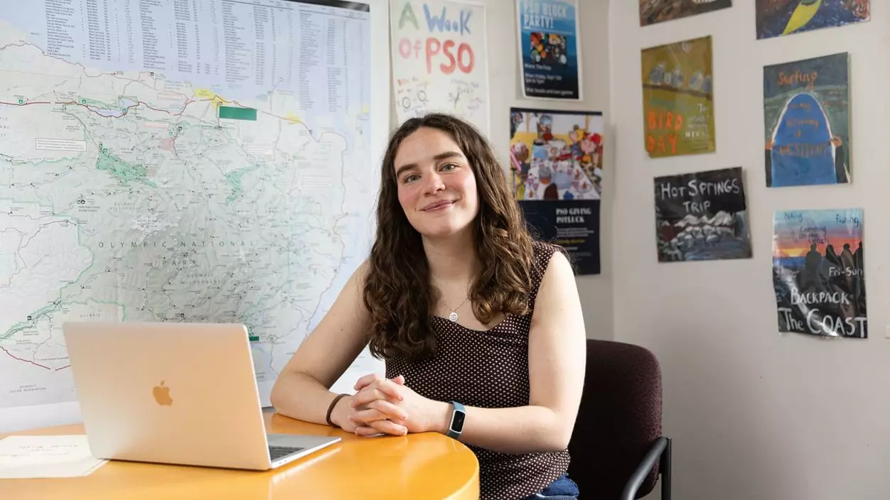 Claire Bargman ’26 at her desk at Puget Sound Outdoors. Behind her are a map of the Olympic Peninsula and flyers for past PSO trips.
