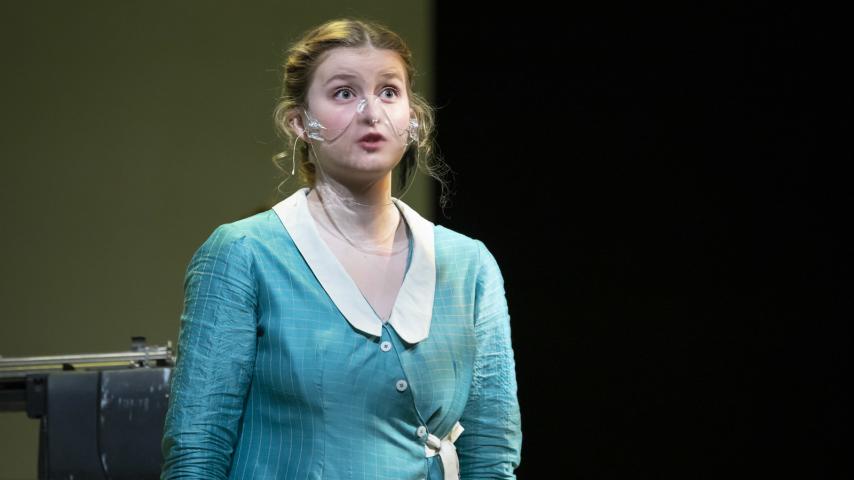 Female student in a teal dress stands onstage in Norton Clapp Theatre in a clear face mask during a performance of Machinal