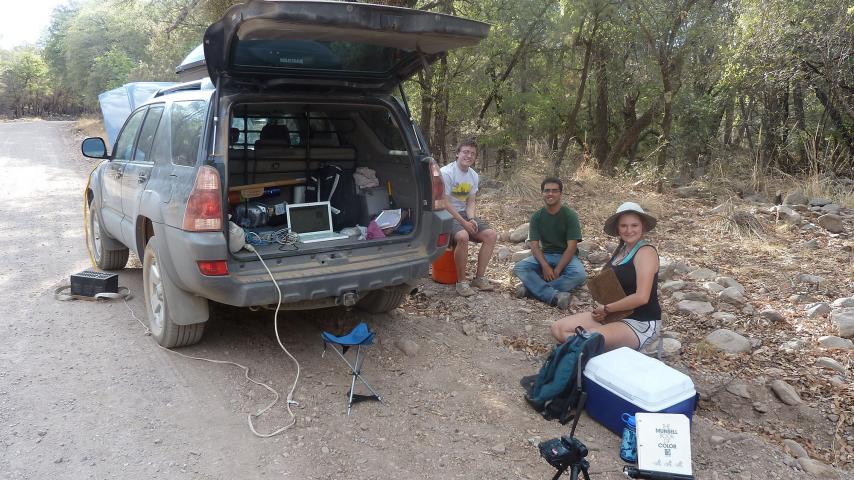 Students sit by an SUV with equipment surrounding them, while conducting fieldwork