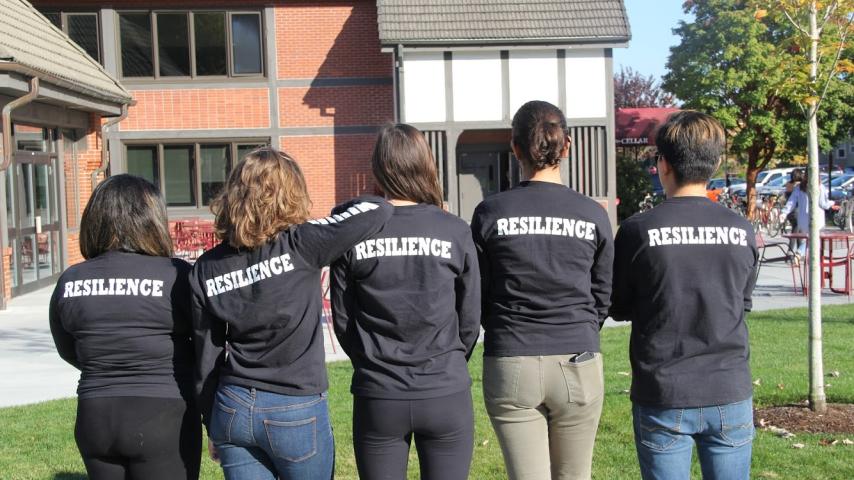 5 students lined up backs to the camera, wearing black shirts that say resilience on the back