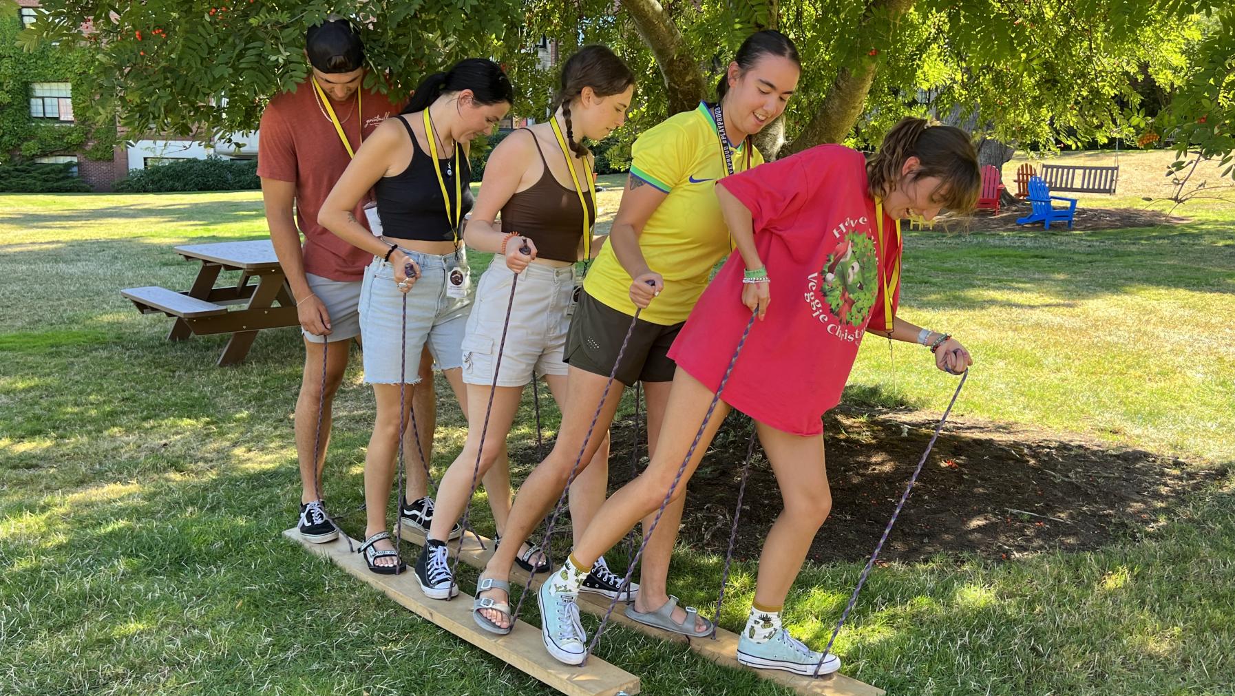 Students stand with either foot on long pieces of wood and hold ropes that attach to the wood planks trying to walk together across a field.