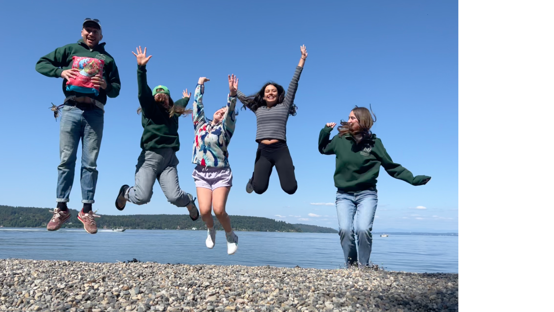 5 students jump into the air with the Puget Sound behind them.