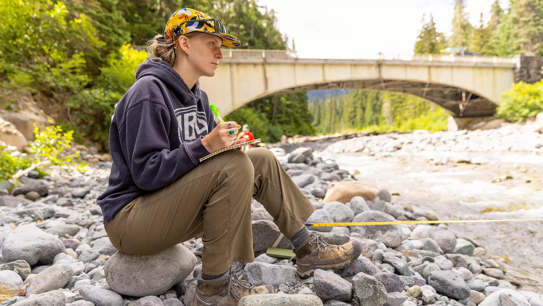 A student sitting on rocks by a river with a notebook.
