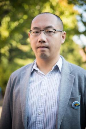 A photo of Professor Sam Liao taken outside. There is a tree in the background, professor Liao is standing wearing a striped shirt, grey blazer, and a button reading He/They