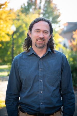 A photo of Professor Justin Tiehen taken outside Wyatt Hall. Justin Tiehen is standing, smiling, and wearing a blue button down shirt with trees visible behind him