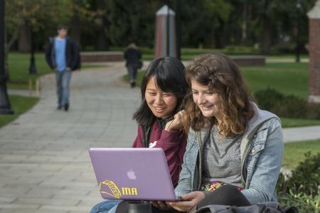 Two female students work on a laptop with Commencement Walk and the Color Post in the background