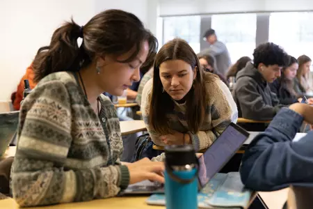 A female student leans over to get a look at another students' laptop