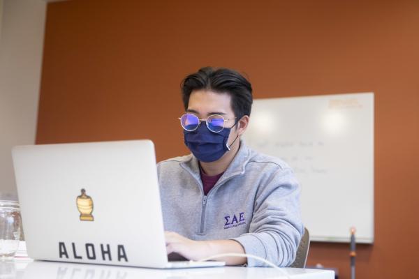 A male student wearing a mask works on his laptop